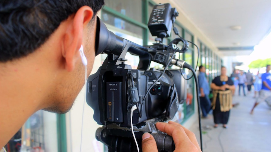 A man looks through the viewfinder of a broadcast quality video camera. woman holding a microphone in the background.