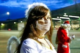A young woman in a face mask and angel costume stands in the foreground while a man in a Santa costume is behind her