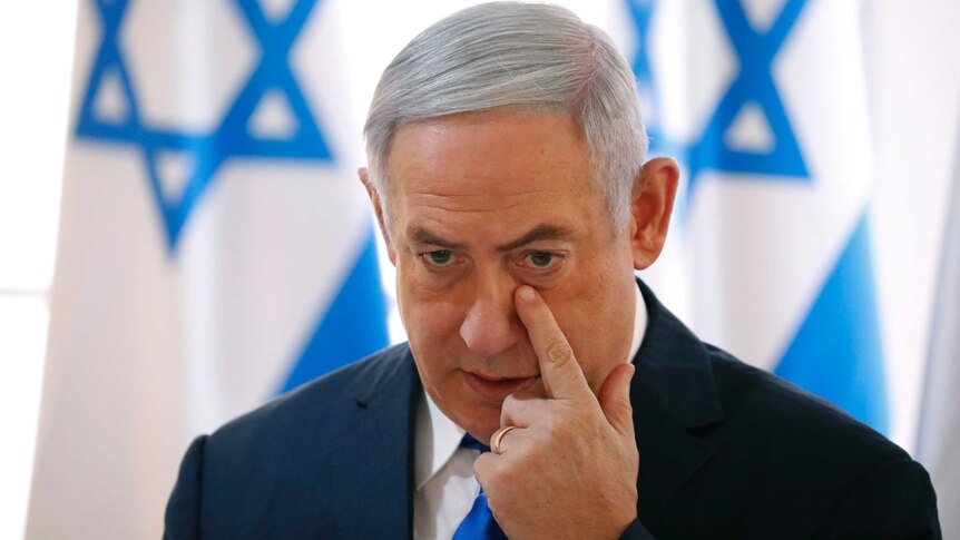 Head and shoulders shot of Benjamin Netanyahu wiping his face with Israeli flags in the background