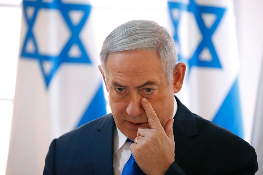 Head and shoulders shot of Benjamin Netanyahu wiping his face with Israeli flags in the background