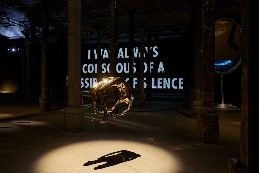 A gold-plated bronze sculpture of a headless body is suspended in the air in a darkened gallery space. 