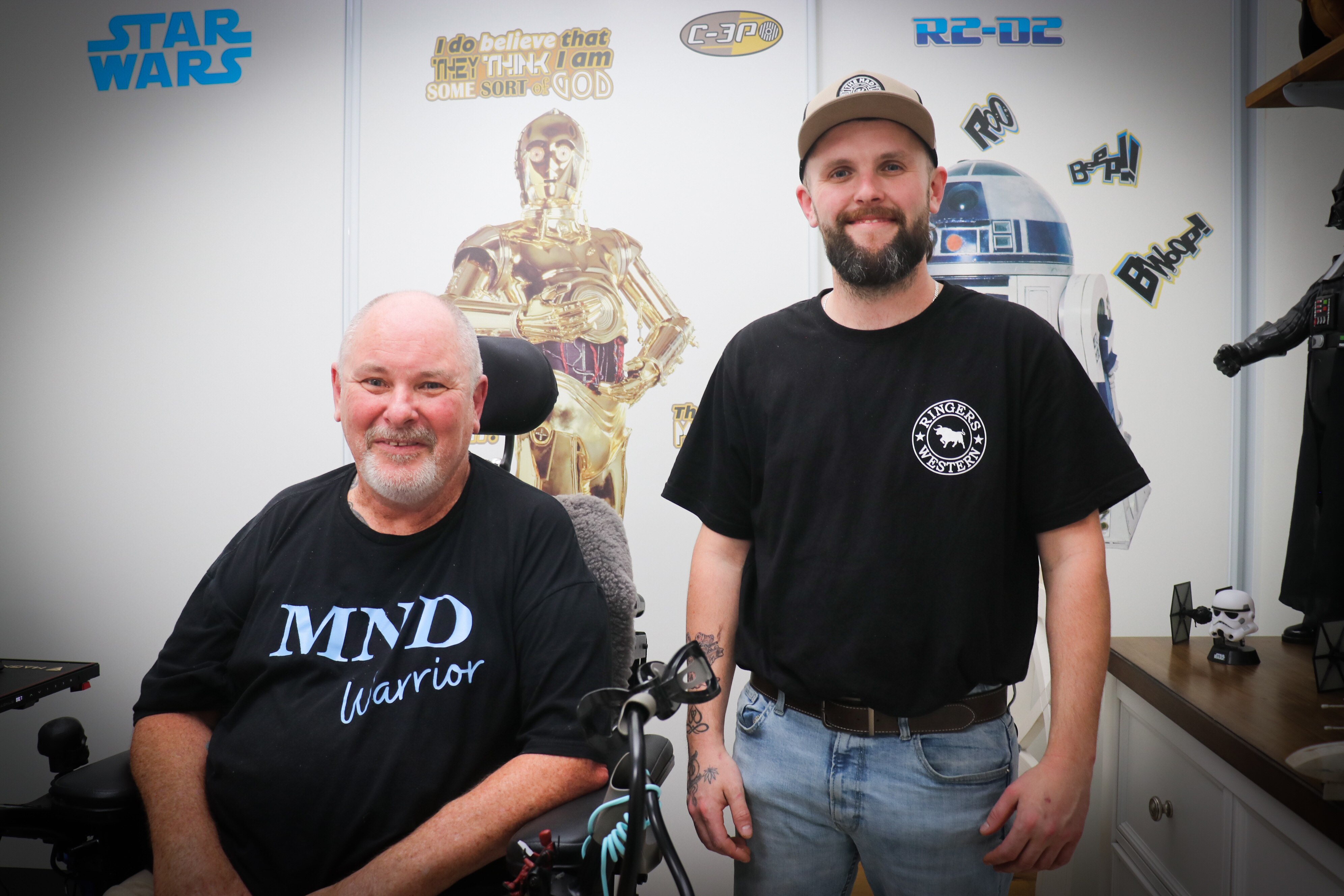 A bald man sitting in a wheelchair pictured with a younger man with a beard smiling at the camera