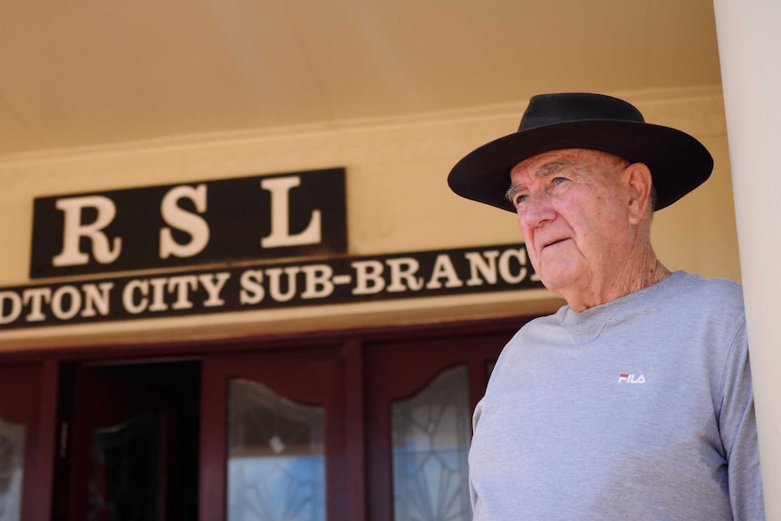 A man wearing an Akubra-style hat stands outside an RSL building, gazing into the distance.