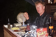 A man wearing a hat sits at an outdoors table surrounded by food and drink.