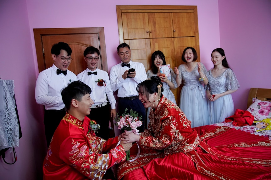 A couple in red wedding clothes in a room with several bridesmaids and groomsmen. 