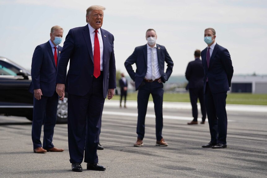An overweight and elderly man in a blue suit stands in front of four suited men wearing face masks