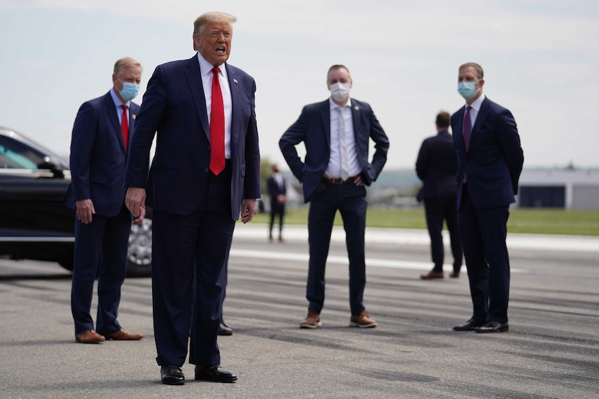 Donald Trump stands in front of four suited men wearing face masks