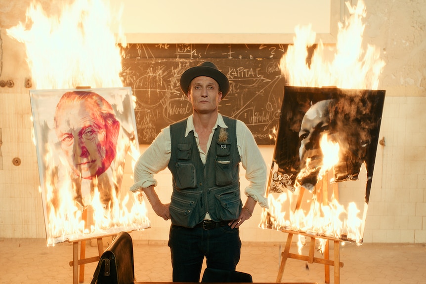 Colour still of Oliver Masucci standing between two burning paintings in white room in 2018 film Never Look Away.