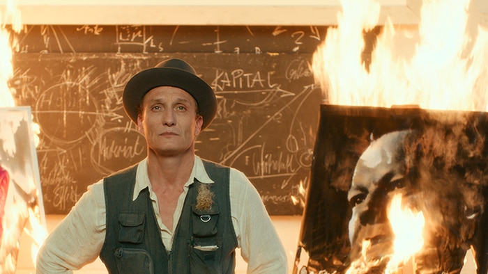 Colour still of Oliver Masucci standing between two burning paintings in white room in 2018 film Never Look Away.