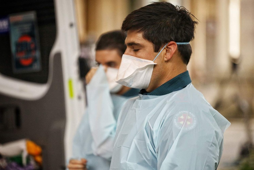 Two Queensland paramedics wearing masks and gowns PPE stand at the back of an ambulance.