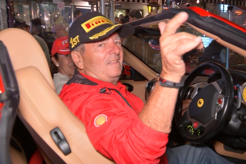 A man wearing a red jacket and black cap sits in the driver's seat of a Ferrari.