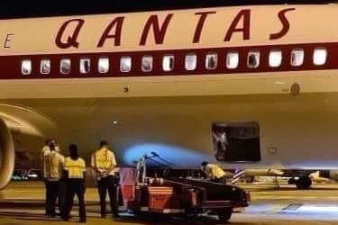 A plane with Qantas logo on the side, on the tarmac of an airport, with ground staff and a vehicle below it.