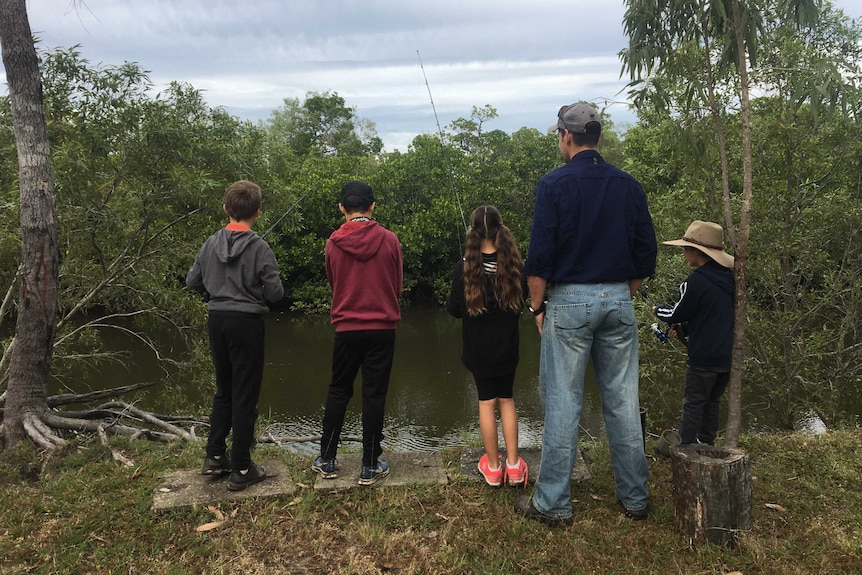 Four kids and a man fish or watch the river. They are standing, surrounded by trees, on an overcast day.
