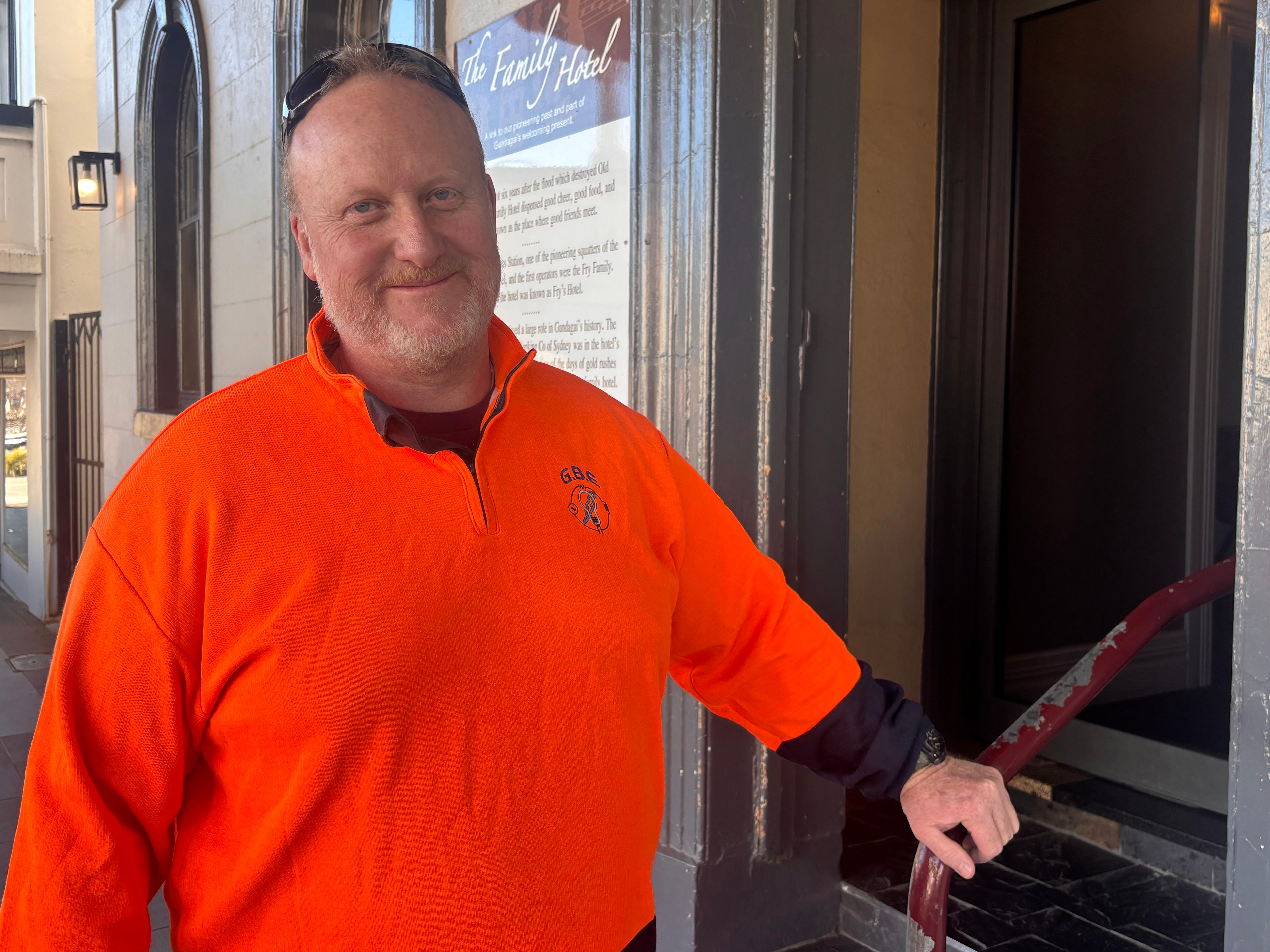 Man in a fluro orange jumper and sunglasses on his head poses in front of the steps of a pub