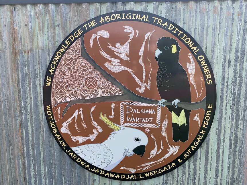 A metal sign on a fence depicting a brown circle with cockatoo motifs and local Indigenous group names around the outside 