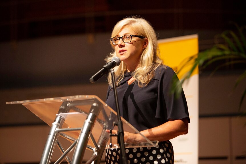 A blonde woman with glasses speaking at a lectern in Newcastle City Hall. She is wearing a black top and polka dot skirt