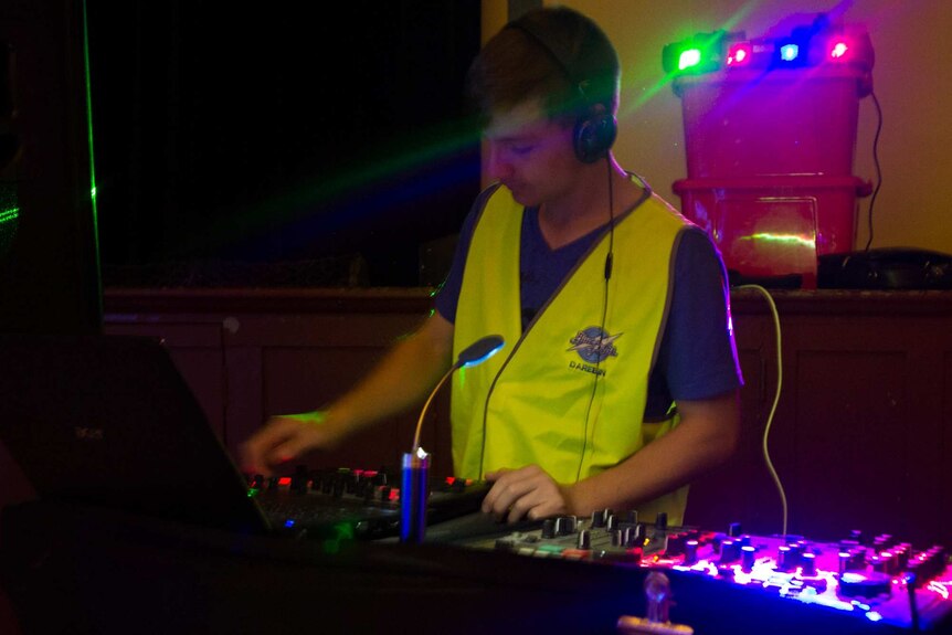 A young man in a high-vis vest works DJ equipment, coloured lights in background.