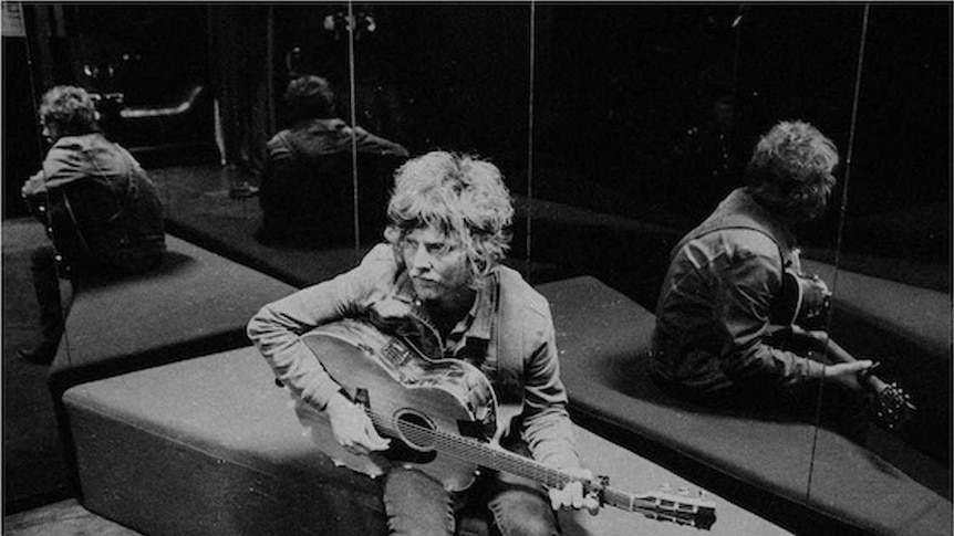 Black and White. Kevin Mitchell is seated with an acoustic guitar. There are mirrors to his back and side, reflecting him.