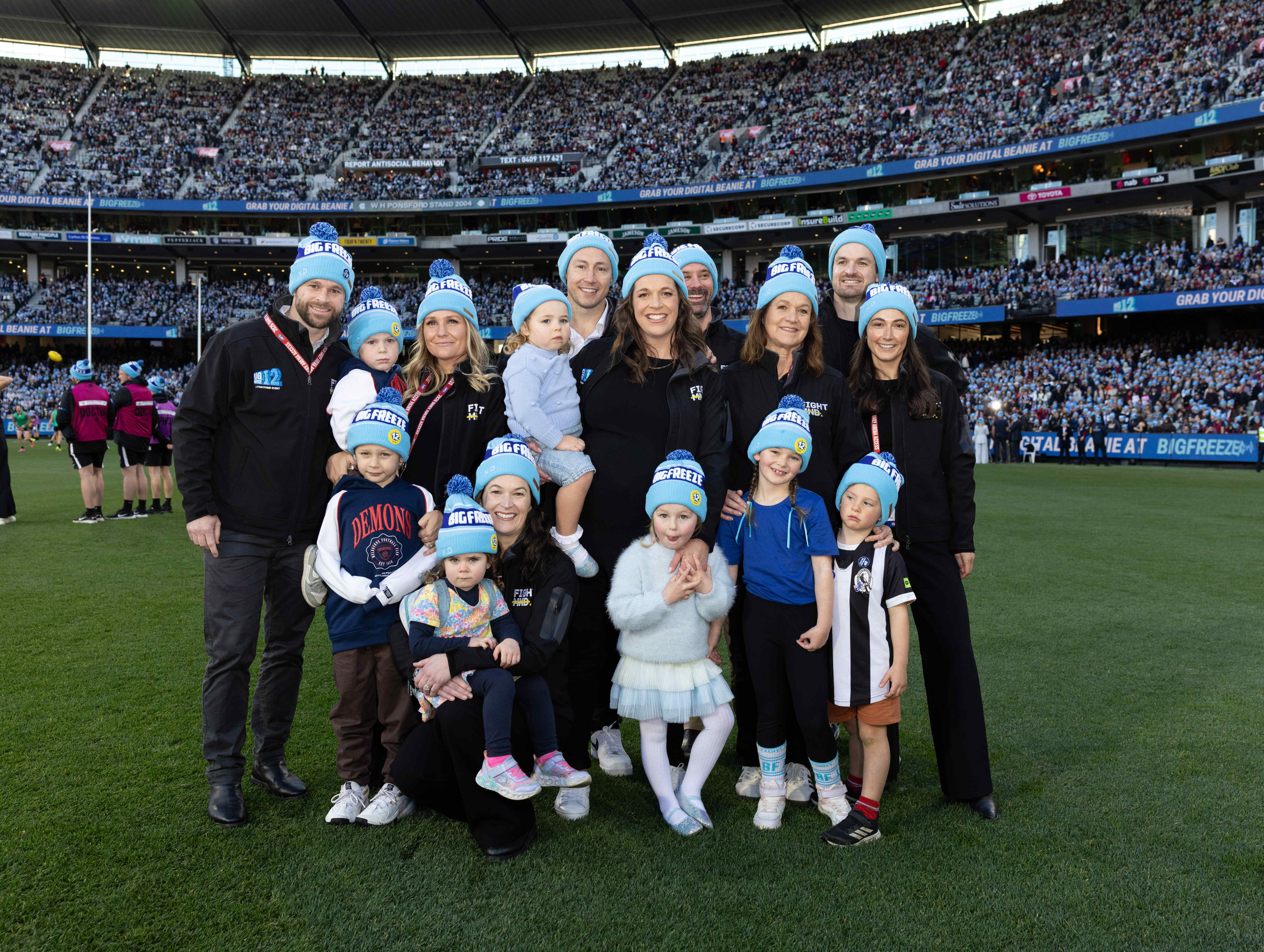 Men, women and children, all wearing blue two-toned beanies and scarves, stand on the grass in a packed sports stadium.