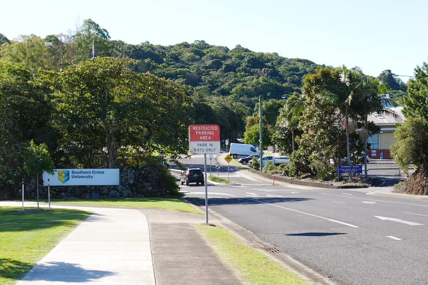 A car drives past a sign reading Southern Cross University. There is a tree-covered hill in the background.