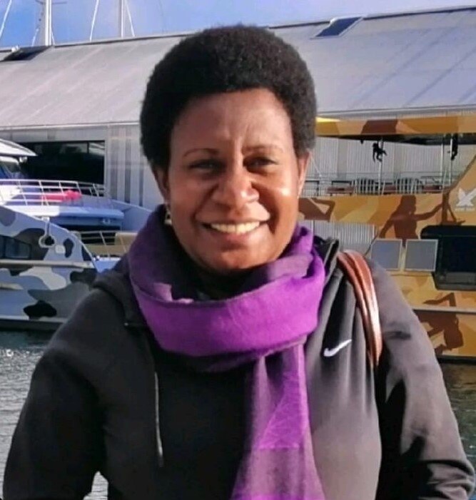 A woman with short dark curly hair wearing a purple scarf and black jumper standing on a jetty on a bright day