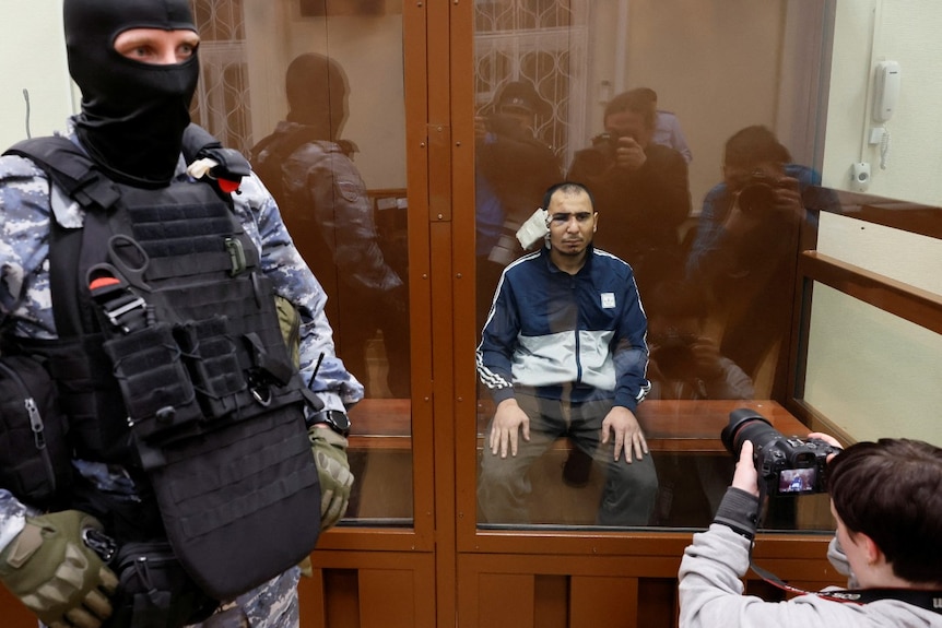 A man with a bandage on his ear sits in a glass box, as an armed guard stands nearby and a journalist takes photographs