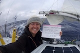 Lisa Blair holds a sign reading HAPPY INTERNATIONAL WOMEN'S DAY sitting on her boat at sea.