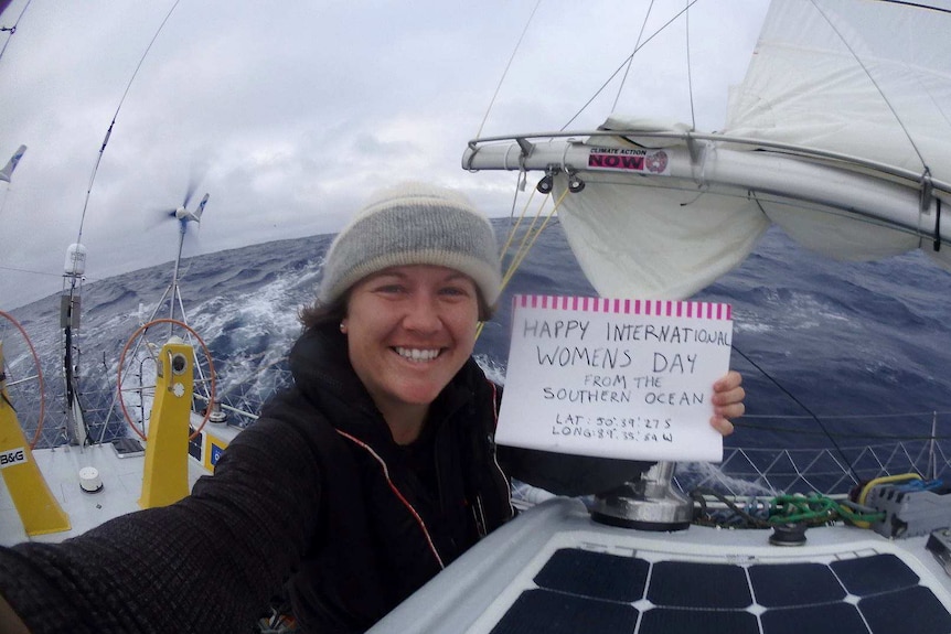 Lisa Blair holds a sign reading HAPPY INTERNATIONAL WOMEN'S DAY sitting on her boat at sea.