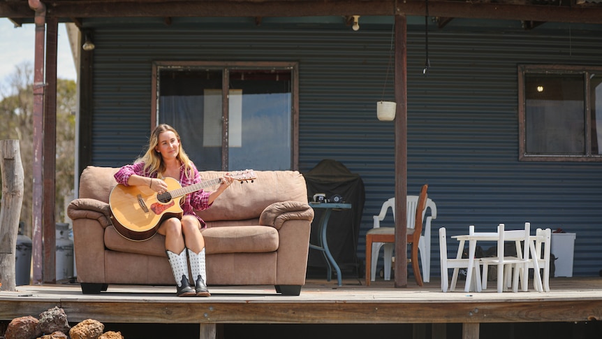 A young blonde woman in a red flannel and cowboy boots plays guitar on the porch of a rustic farmhouse.