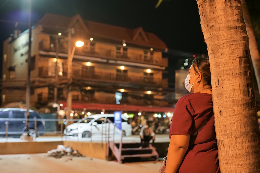 A woman wearing a mask and a red shirt looks over a house sitting on a street and lit up by a lamp.