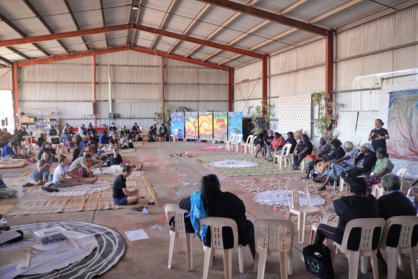 A wide shot of the shed with dozens of women sitting amongst artwork on the floor.
