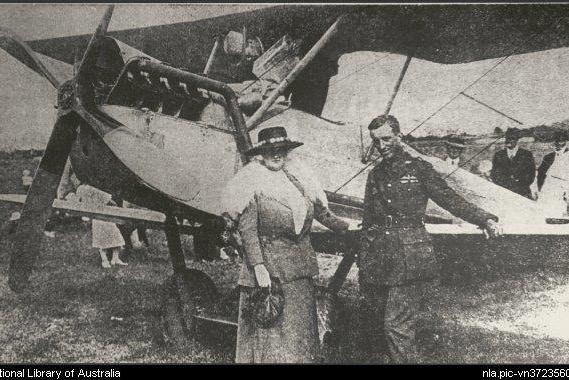 Scratchy monochrome photo of a well-dressed woman and Lieutenant Long in front of a biplane on the ground.