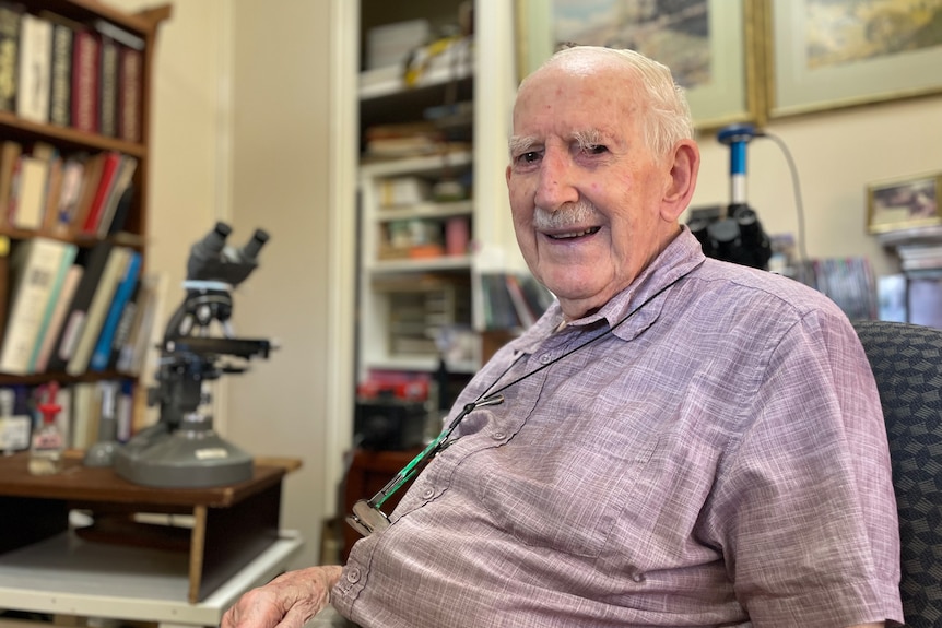 An old man sits at a desk in front of a microscope 