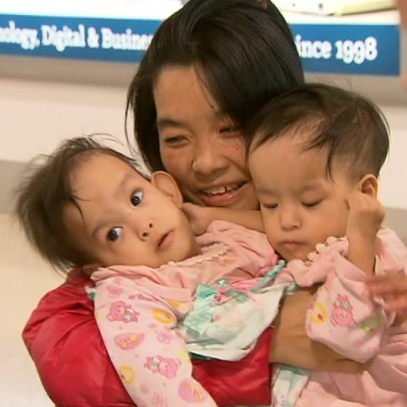 Bhumchu Zangmo smiles at her twin daughters who she is cradling on her lap while sitting in a wheelchair at the airport.