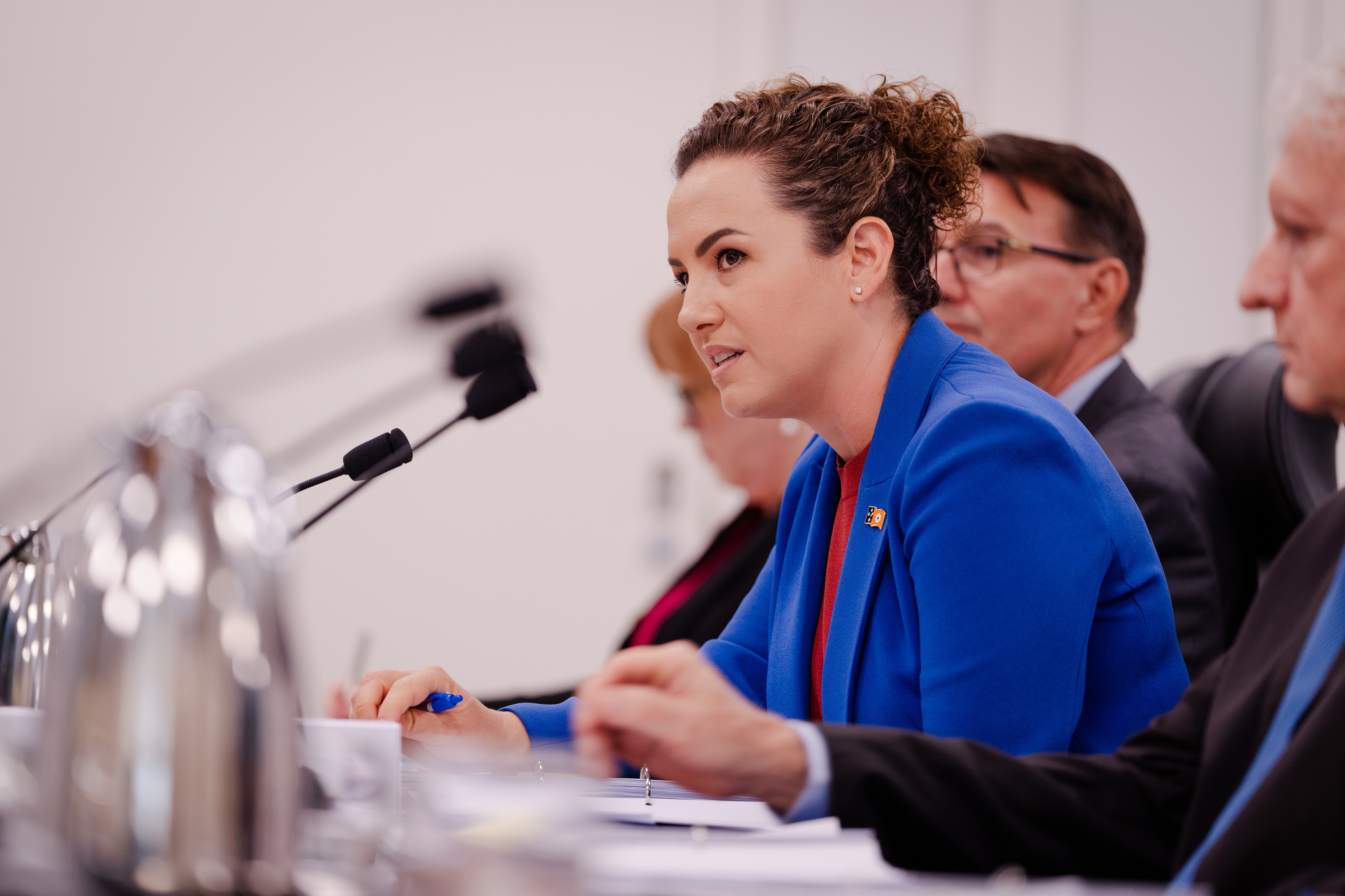 A white woman with brown curly hair tied up in bun, wearing blue blazer sitting at desk with others, serious expression.