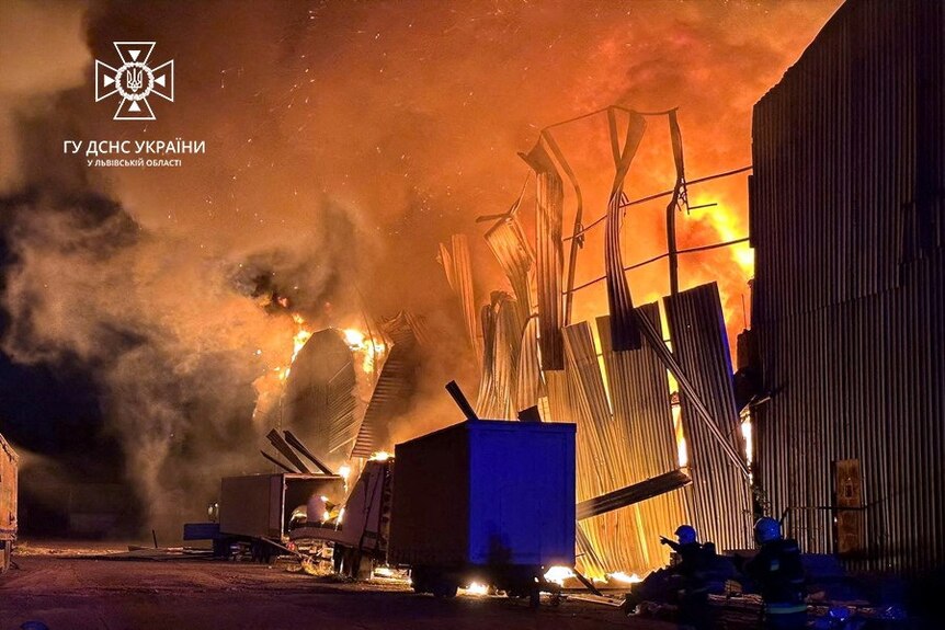 Bright orange flames engulf a warehouse, with two trucks parked on the road beside it.