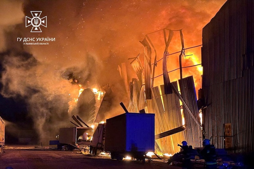 Bright orange flames engulf a warehouse, with two trucks parked on the road beside it.