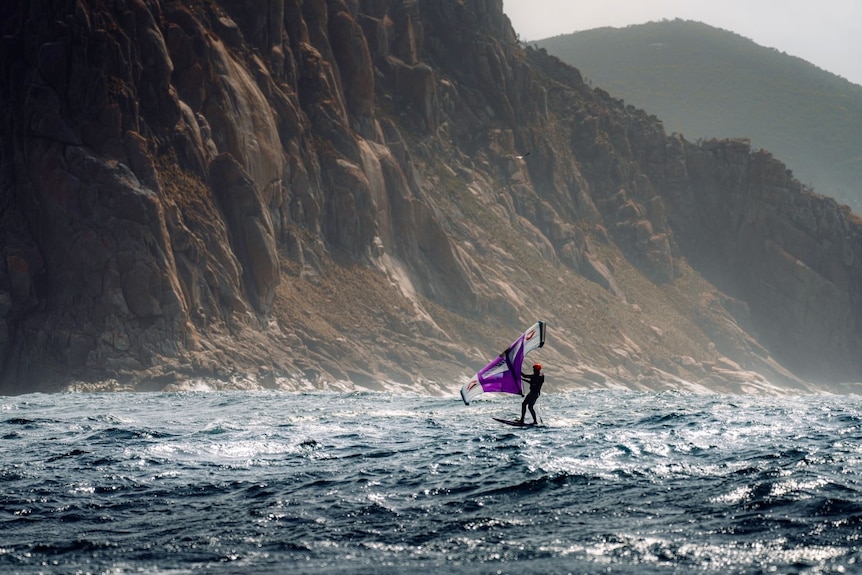 A man on a windsurfer seen on the ocean with cliffs in the background.