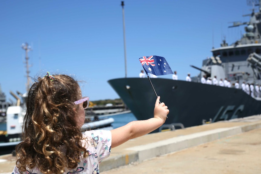 A girl wearing sunglasses and waving an Australian flag stands on a dock looking towards a navy ship