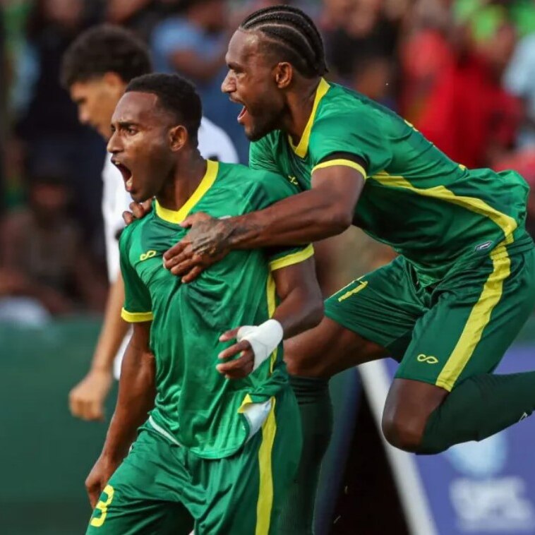 Two Vanuatu soccer players wearing green and yellow trim jerseys jump high in celebration on pitch
