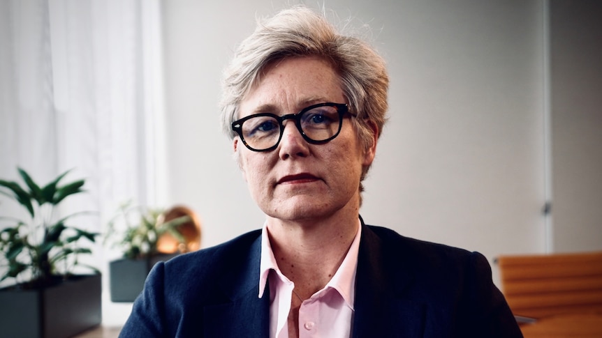 A woman with short grey hair and glasses, wearing a dark suit over a light pink shirt looks into the camera.