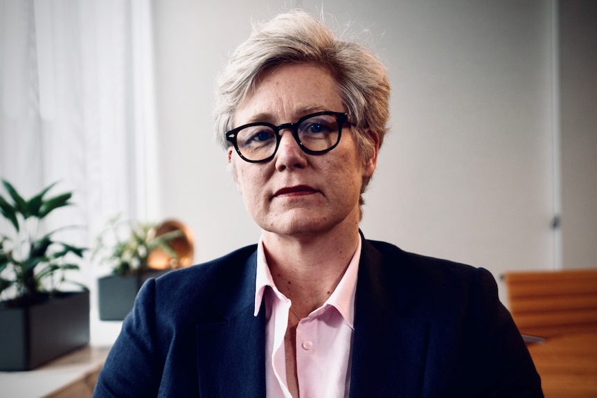 A woman with short grey hair and glasses, wearing a dark suit over a light pink shirt looks into the camera.