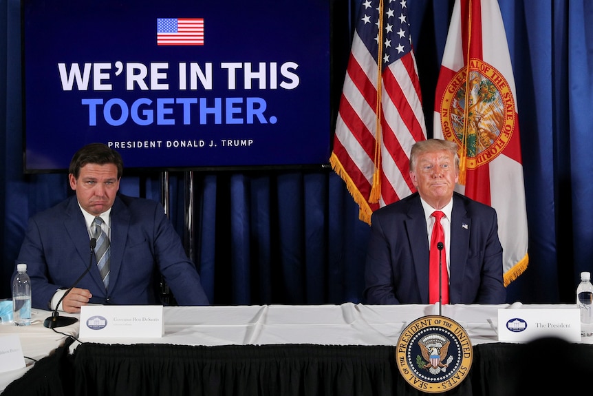 Donald Trump and Ron DeSantis sit behind a table frowning in front of a sign that reads "we're in this together"
