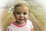 A blonde baby girl sits on a deck, smiling at the camera and wearing a flower in her hair.
