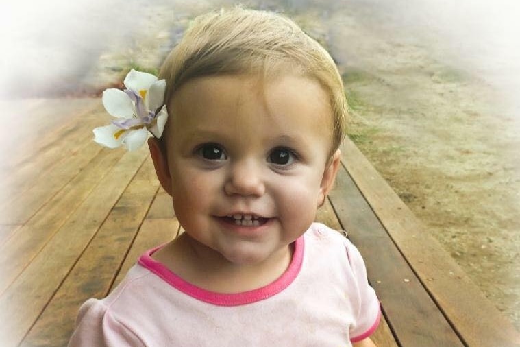A blonde baby girl sits on a deck, smiling at the camera and wearing a flower in her hair.