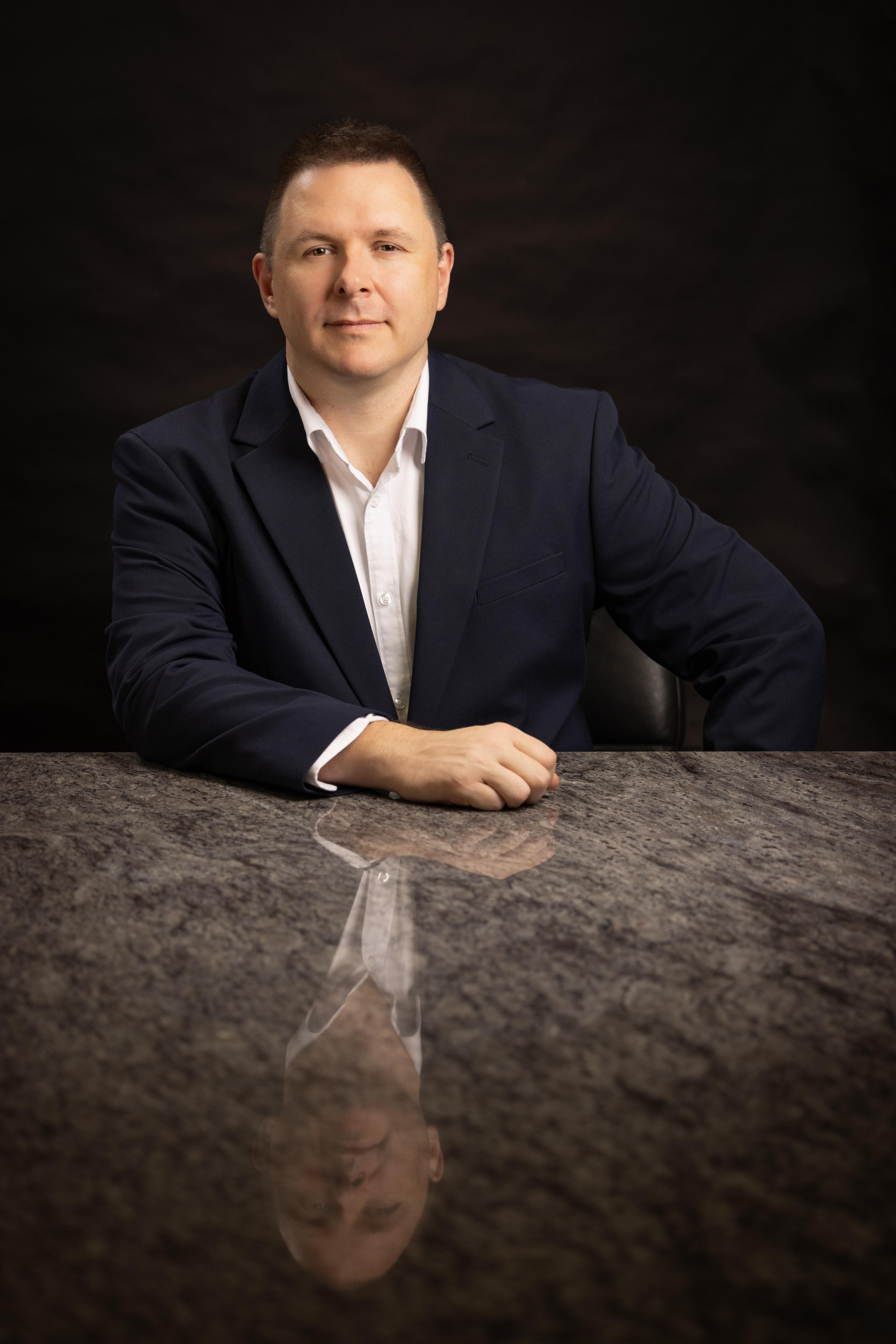 A middle-aged man in a suit poses for a photo with his hands in his pockets.