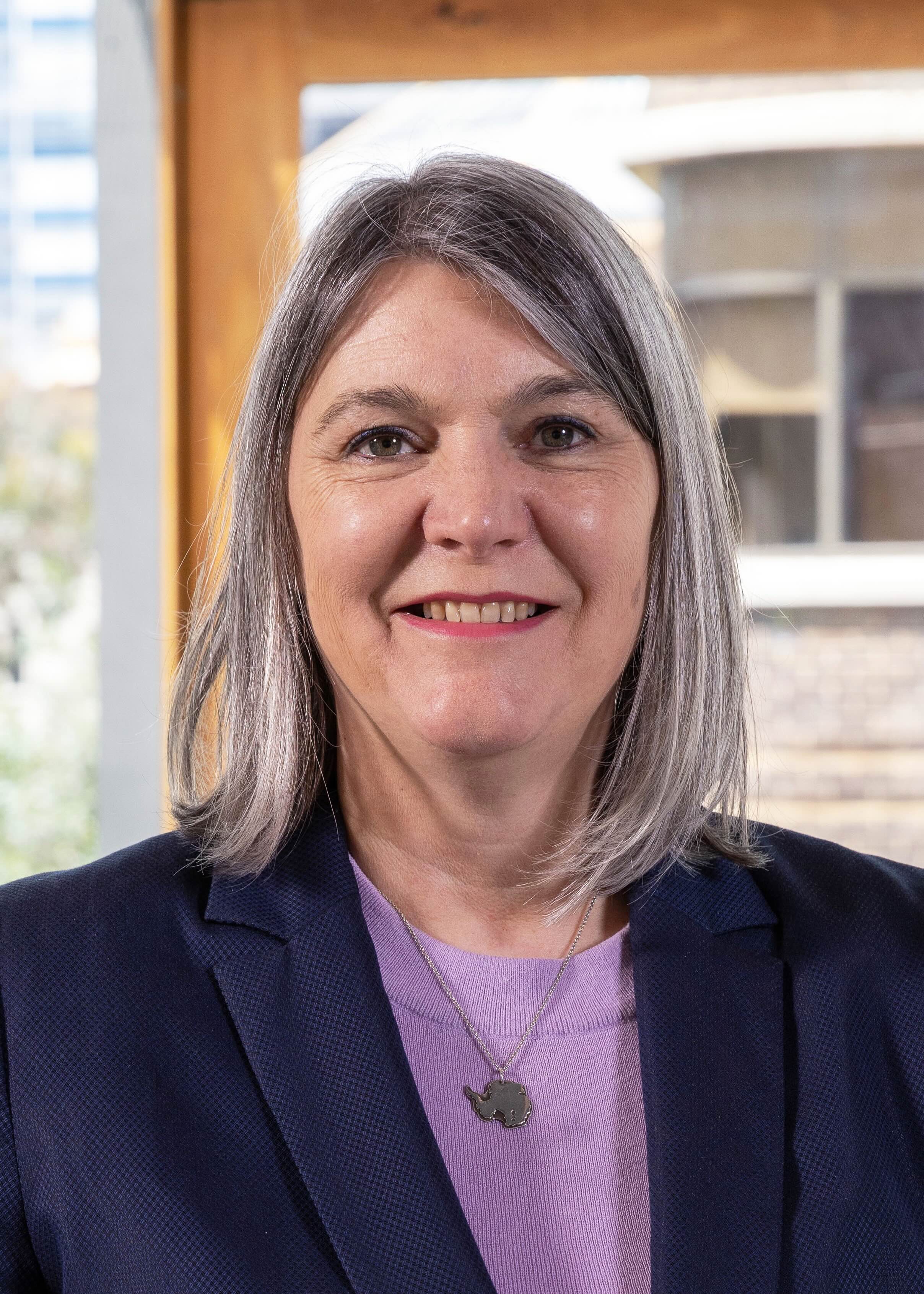 Mulher de camisa roxa e jaqueta sorri para a câmera.