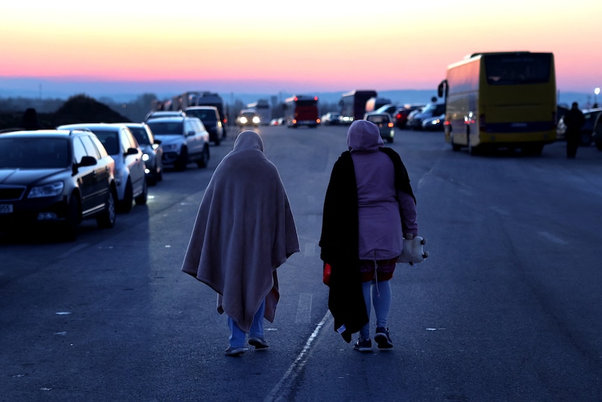 Two women wrapped in blankets walk away from the camera down a freeway with cars gridlocked in both directions.