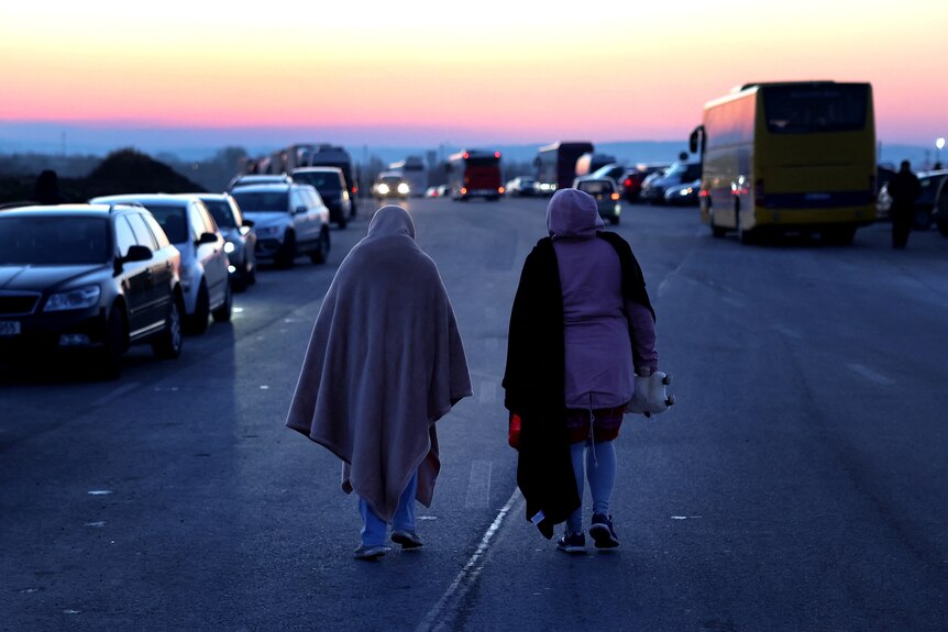Due donne avvolte in coperte si allontanano dalla telecamera su un'autostrada con le auto bloccate in entrambe le direzioni.