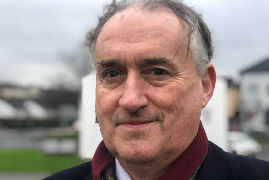 Eamonn McDermott smiles in headshot against grey sky with green grass. He wears maroon paisley scarf and navy coat.